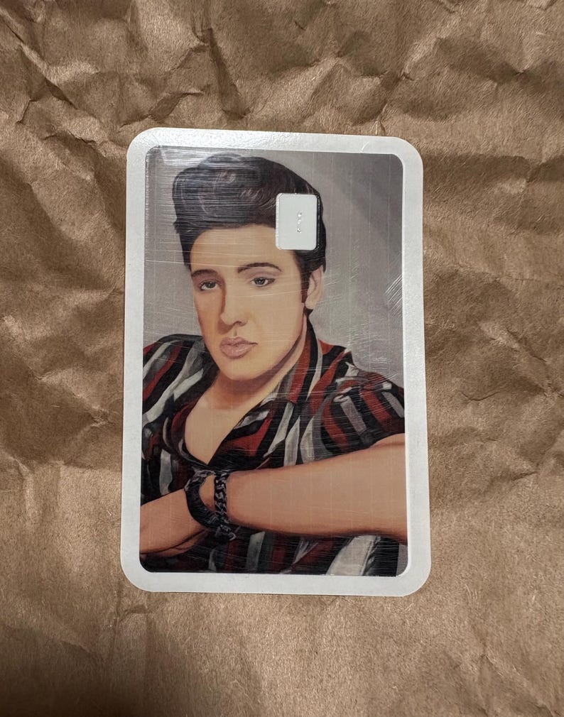 May include: A rectangular card featuring a portrait of a man with dark hair and a striped shirt in shades of red, black, and white. The man is wearing a black bracelet. The card has a white border.