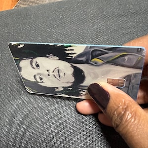 May include: A rectangular card featuring a portrait of a person with long dreadlocks. The card has a light blue border and a gold-colored chip. The person's face is the focal point of the image.