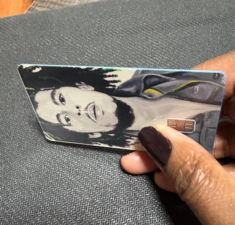 May include: A rectangular card featuring a portrait of a person with long dreadlocks. The card has a light blue border and a gold-colored chip. The person's face is the focal point of the image.