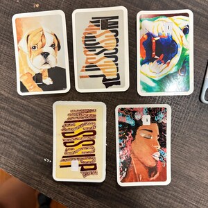 May include: Five rectangular decorative items with white borders. The images include a bulldog, abstract patterns, a colorful dog, a pattern with the word "Mississippi", and a portrait of a woman with colorful hair.