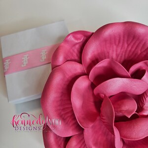 K&ouml;nnte beinhalten: Eine gro&szlig;e, leuchtend pinkfarbene Stoffblume mit mehreren Bl&uuml;tenbl&auml;ttern. Die Blume ist scharf, w&auml;hrend eine wei&szlig;e Schachtel mit pinkfarbenem Band im Hintergrund unscharf ist.