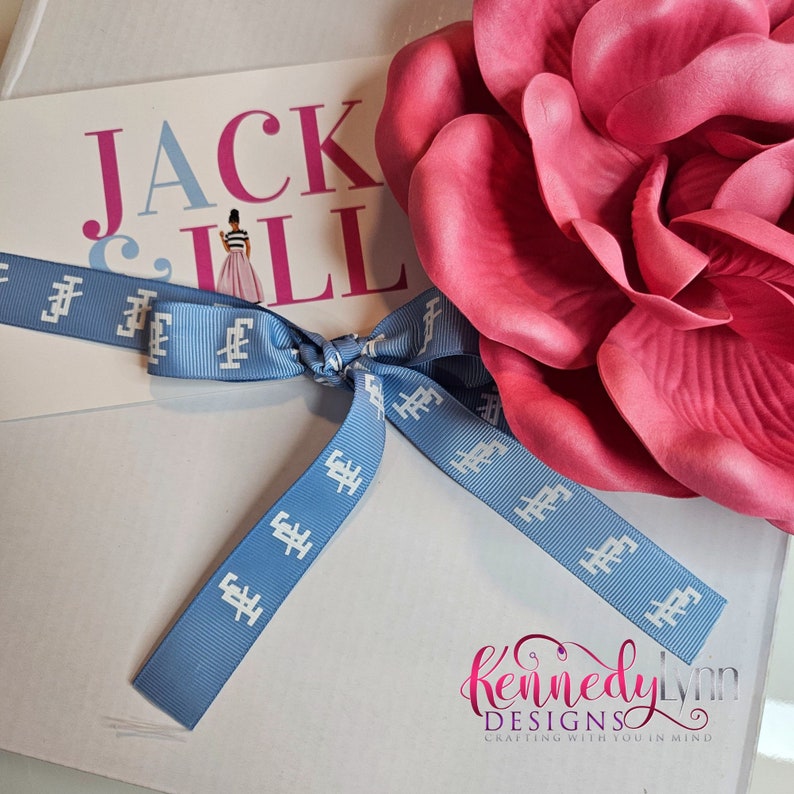 K&ouml;nnte beinhalten: Eine wei&szlig;e Geschenkbox mit einem blauen Band, das zu einer Schleife gebunden ist. Das Band hat wei&szlig;en Text mit der Aufschrift "Jack & Jill". Eine gro&szlig;e rosa Blume liegt auf der Box. Die Box ist mit "KennedyLynn Designs, Crafting with you in mind." beschriftet.