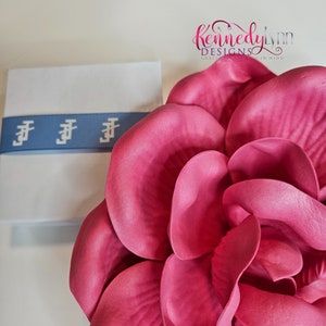 K&ouml;nnte beinhalten: Eine gro&szlig;e, rosafarbene Stoffblume mit mehreren Lagen Bl&uuml;tenbl&auml;tter. Die Blume liegt auf einer wei&szlig;en Oberfl&auml;che neben einer kleinen wei&szlig;en Schachtel mit einem blauen Band.