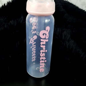 Könnte beinhalten: Eine klare Babyflasche mit einem rosa Deckel und einem rosa Vinyl-Aufkleber mit der Aufschrift "Christine" und "Princess".