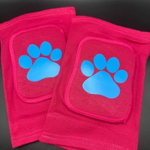 May include: A pair of pink knee pads with blue paw print designs. The knee pads are made of a soft, stretchy fabric and have a padded section for protection.