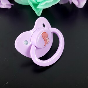 May include: A purple pacifier with a round, white nipple and a pink dinosaur design on the front.