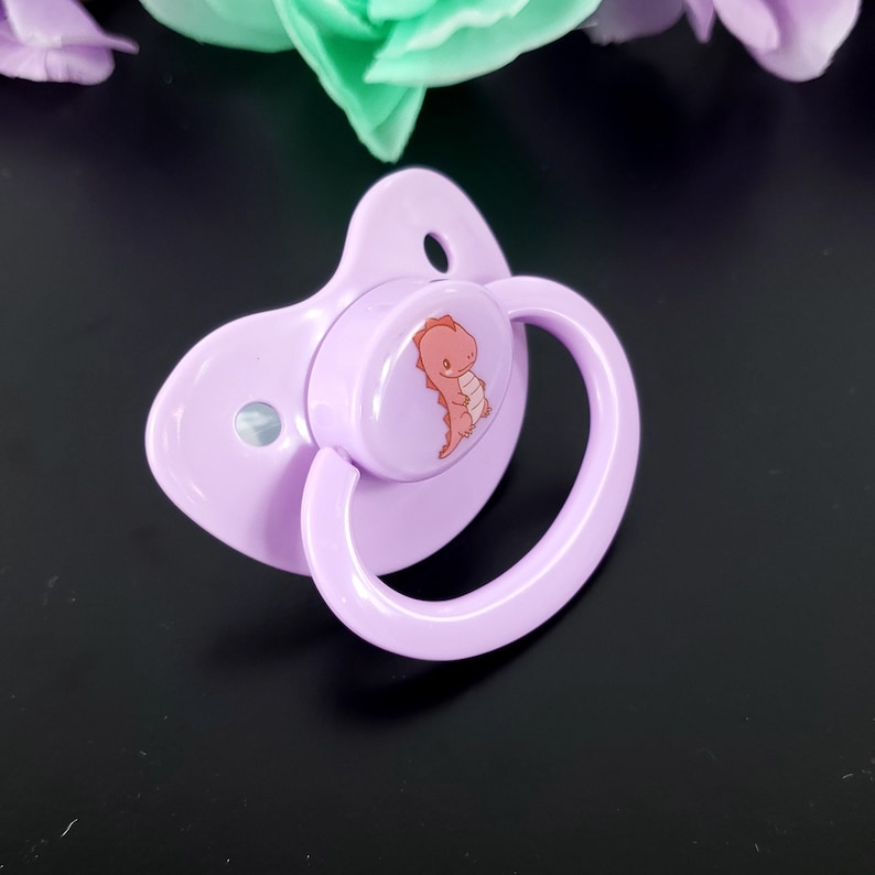 May include: A purple pacifier with a round, white nipple and a pink dinosaur design on the front.