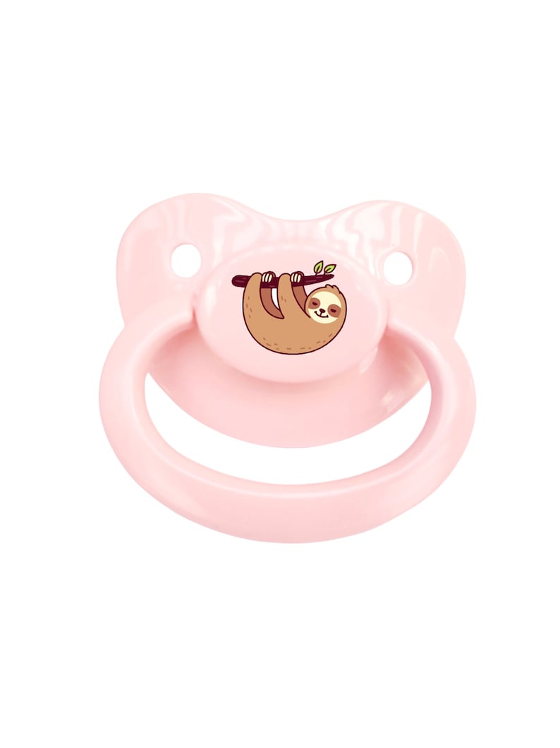 May include: A pink pacifier with a sloth design. The sloth is brown and hanging from a branch with green leaves.