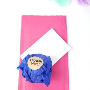 May include: A pink padded envelope with a white card and a blue tissue paper wrapped package with a brown sticker that says "THANK YOU" with a red heart.