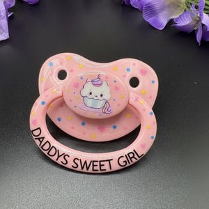 May include: Pink pacifier with a cupcake design and the text "DADDYS SWEET GIRL". The pacifier has a round handle and a flat, shield-shaped front with polka dots and heart accents. The cupcake has a smiling face and a purple swirl on top.