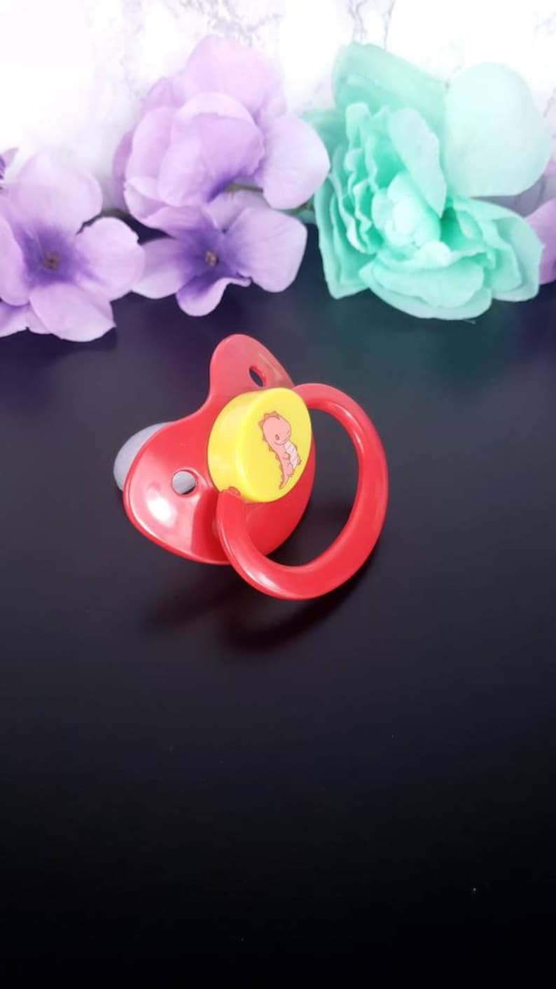 May include: A red pacifier with a yellow and white dinosaur design. The pacifier has a ring handle.