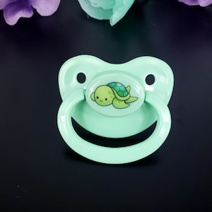 Könnte beinhalten: Ein hellgrüner Schnuller mit einem Schildkröten-Design. Die Schildkröte ist grün mit einer braunen Schale und einem lächelnden Gesicht.