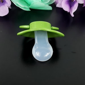 May include: A green silicone pacifier with a white silicone nipple. The pacifier has a flat, circular base with two small handles on the sides.