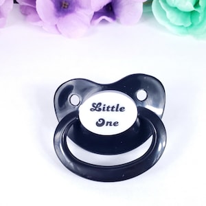 Puede incluir: Chupón negro con un inserto blanco ovalado que dice "Little One" en escritura negra.