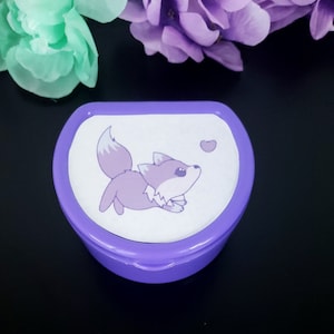 May include: A small purple plastic container with a white lid featuring a cartoon purple fox with a white tail and a pink heart. The container is designed to hold small items like jewelry or medication.