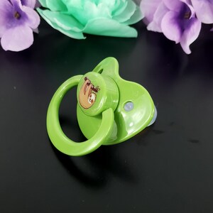 May include: A green pacifier with a round, white nipple and a green ring. The pacifier has a round, green disc with a cartoon image of a brown sloth hanging from a branch.