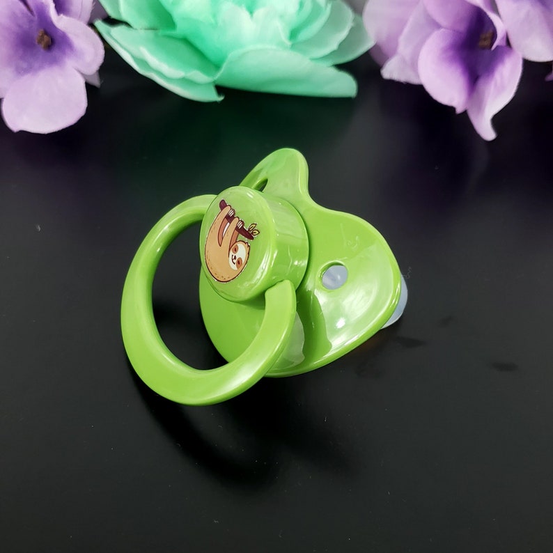 May include: A green pacifier with a round, white nipple and a green ring. The pacifier has a round, green disc with a cartoon image of a brown sloth hanging from a branch.