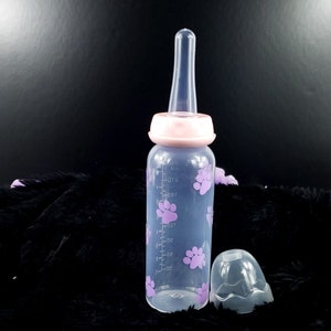 May include: A clear plastic baby bottle with a pink lid and purple paw prints. The bottle has markings for measuring ounces. The bottle comes with a separate clear plastic nipple.