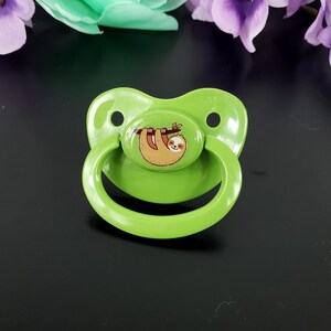 May include: A green pacifier with a sloth design. The sloth is brown and hanging from a branch. The pacifier has a round, flat nipple and a handle.