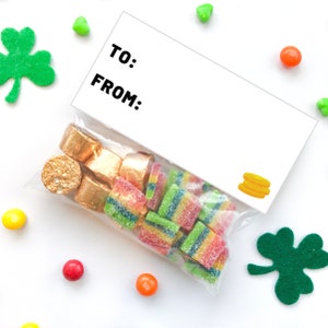 Puede incluir: Una bolsa de regalo blanca con un gr&aacute;fico de moneda de oro y una etiqueta que dice "Para:" y "De:". La bolsa est&aacute; llena de caramelos gomosos coloridos y chocolates envueltos en oro.