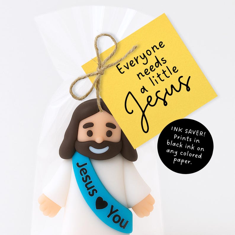 May include: A figurine of Jesus with brown hair and a beard, wearing a white robe and a blue sash that says "Jesus You." The figurine is attached to a yellow tag with the text "Everyone needs a little Jesus."