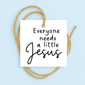 May include: A white square tag with the text "Everyone needs a little Jesus" in black script. The tag is suspended by a tan twine loop against a light blue background.