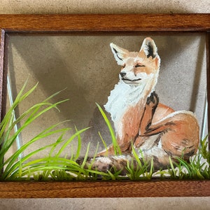 Op de afbeelding: Een ingelijst schilderij van een rode vos die in een veld met groen gras zit. De vos kijkt naar rechts en heeft zijn rechterpoot opgeheven. Het schilderij is in een houten lijst.