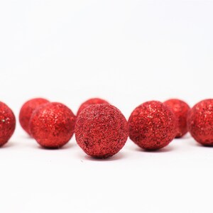 May include: A group of ten small, red, glitter-covered balls. The balls are arranged in a semi-circle on a white background.