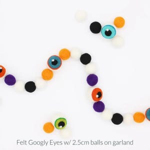 Peut inclure: Une guirlande compos&eacute;e de boules en feutrine de 2,5 cm de couleur orange, noire, blanche et violette, altern&eacute;es avec des yeux mobiles en feutrine bleus, verts et orange. Le texte "Felt Googly Eyes w/ 2.5cm balls on garland" est inscrit en bas.