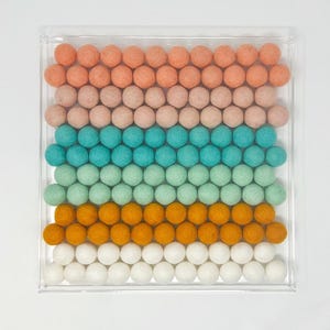 Puede incluir: Una caja de acr&iacute;lico transparente llena de bolas de fieltro de colores. Las bolas est&aacute;n dispuestas en filas, cada fila de un color diferente: melocot&oacute;n, rosa claro, turquesa, verde claro, naranja y blanco.