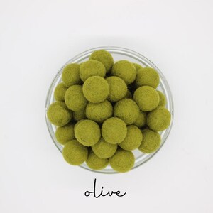 May include: A clear glass bowl filled with olive-green felt balls. The word "olive" is written in a cursive font below the bowl. The felt balls are round and densely packed, creating a textured appearance. The background is plain white.
