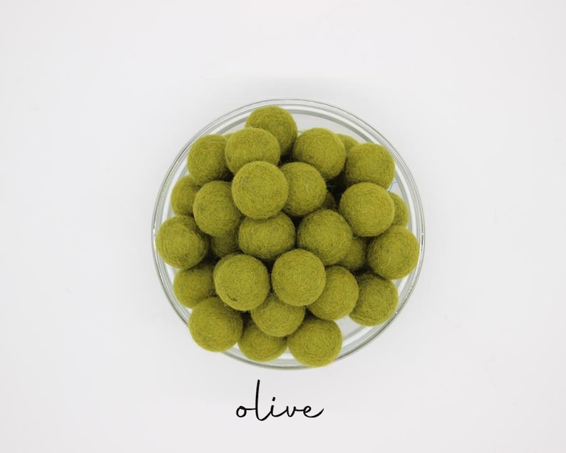 May include: A clear glass bowl filled with olive-green felt balls. The word "olive" is written in a cursive font below the bowl. The felt balls are round and densely packed, creating a textured appearance. The background is plain white.