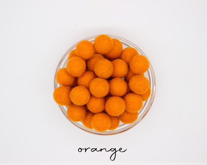 May include: A glass bowl containing many orange felt balls. The word "orange" is written in cursive script below the bowl. The felt balls are a bright, solid colour, and the bowl sits on a white surface.