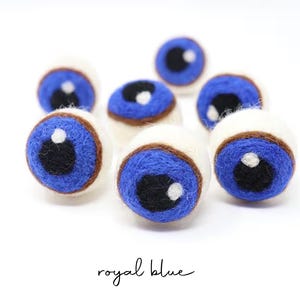 Peut inclure: Un ensemble de sept boules en laine feutr&eacute;e sph&eacute;riques, chacune pr&eacute;sentant une base blanche, un iris bleu roi, une pupille noire et un petit point blanc brillant. Le texte royal blue est imprim&eacute; en &eacute;criture noire au bas de l'image.