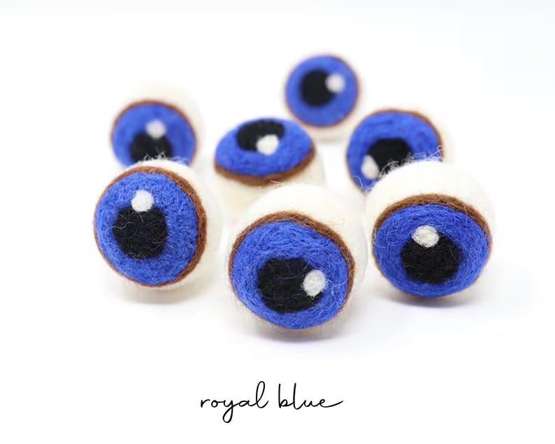Peut inclure: Un ensemble de sept boules en laine feutr&eacute;e sph&eacute;riques, chacune pr&eacute;sentant une base blanche, un iris bleu roi, une pupille noire et un petit point blanc brillant. Le texte royal blue est imprim&eacute; en &eacute;criture noire au bas de l'image.