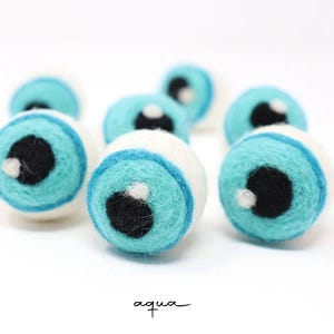Peut inclure: Un ensemble de boules en laine feutr&eacute;e sph&eacute;riques ressemblant &agrave; des globes oculaires. Chaque boule pr&eacute;sente une base blanche, un iris bleu aqua vif, une pupille noire et un petit point blanc brillant. Le texte aqua est inscrit en bas.