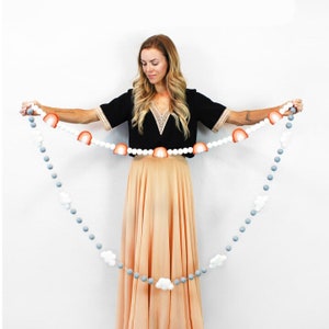 Puede incluir: Una guirnalda de pompones blancos y grises con detalles de arco&iacute;ris y nubes de fieltro. La guirnalda est&aacute; enhebrada en una cuerda blanca y la est&aacute; sujetando una persona.