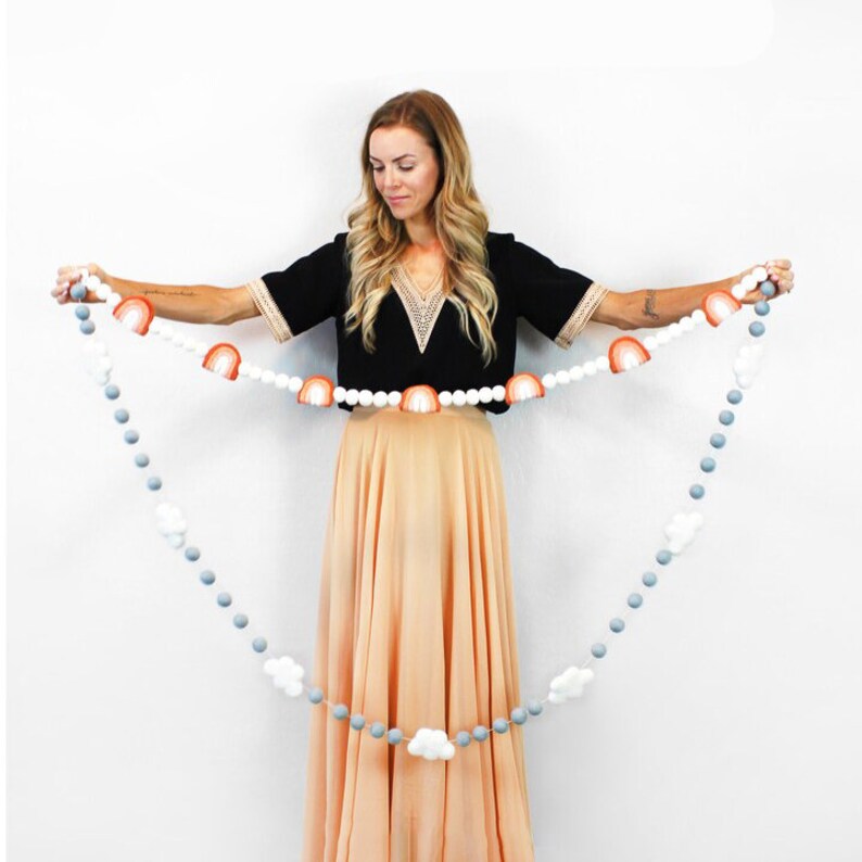 Puede incluir: Una guirnalda de pompones blancos y grises con detalles de arco&iacute;ris y nubes de fieltro. La guirnalda est&aacute; enhebrada en una cuerda blanca y la est&aacute; sujetando una persona.