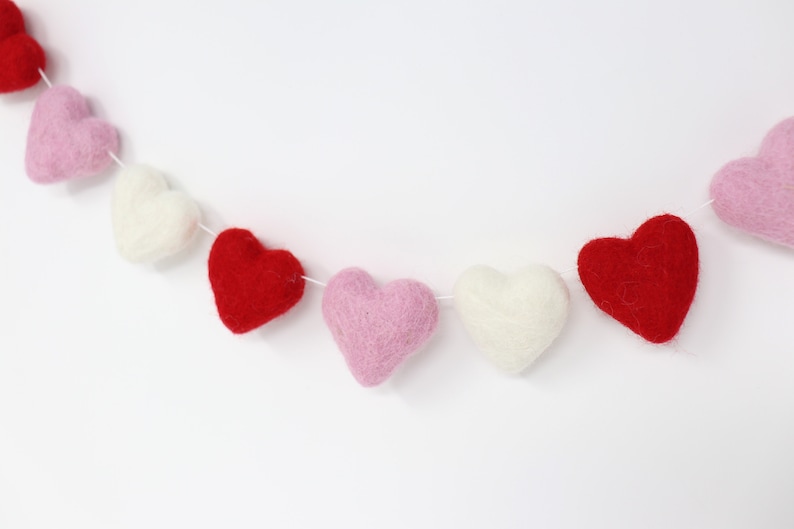 May include: A garland of felt hearts in pink, red, and white. The hearts are strung together with white thread and are arranged in a repeating pattern.
