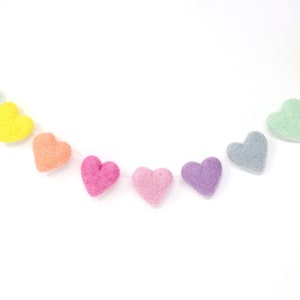 May include: A colorful garland of felt hearts in shades of pink, purple, orange, yellow, green, and gray. The hearts are strung together on a white string, creating a decorative banner. Perfect for Valentine's Day or other celebrations.