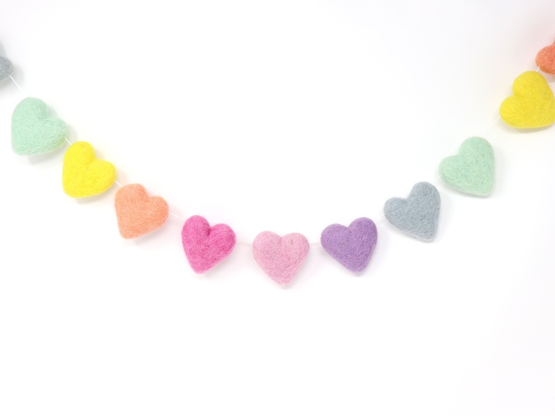 May include: A colorful garland of felt hearts in shades of pink, purple, orange, yellow, green, and gray. The hearts are strung together on a white string, creating a decorative banner. Perfect for Valentine's Day or other celebrations.