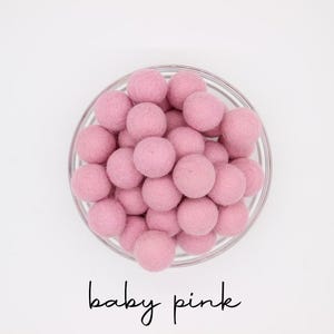 May include: A glass bowl filled with baby pink felt balls. The balls are round and have a soft, textured appearance. The words "baby pink" are written in a cursive font below the bowl.