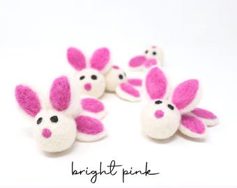 May include: A collection of small, handcrafted felted wool bunny figures. Each bunny features a round white head with bright pink ears, black felt eyes, and a small pink nose. The text bright pink is written in black script below the figures.