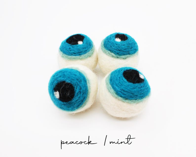 Peut inclure: Quatre boules en laine feutr&eacute;e de forme sph&eacute;rique dispos&eacute;es en carr&eacute;. Chaque boule pr&eacute;sente une base blanc cass&eacute; avec un motif superpos&eacute; vert menthe et bleu paon, surmont&eacute; d'un centre noir circulaire avec un petit point blanc. Texte : peacock / mint.