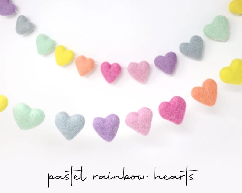 May include: Pastel rainbow heart garlands in shades of pink, purple, yellow, orange, and green. The felt hearts are strung together, creating a cheerful and colorful decoration. The text "pastel rainbow hearts" is written in a script font.