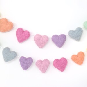 May include: A colorful felt heart garland in pastel shades of yellow, orange, pink, purple, and blue. The hearts are strung together on a white string, creating a festive and decorative item.