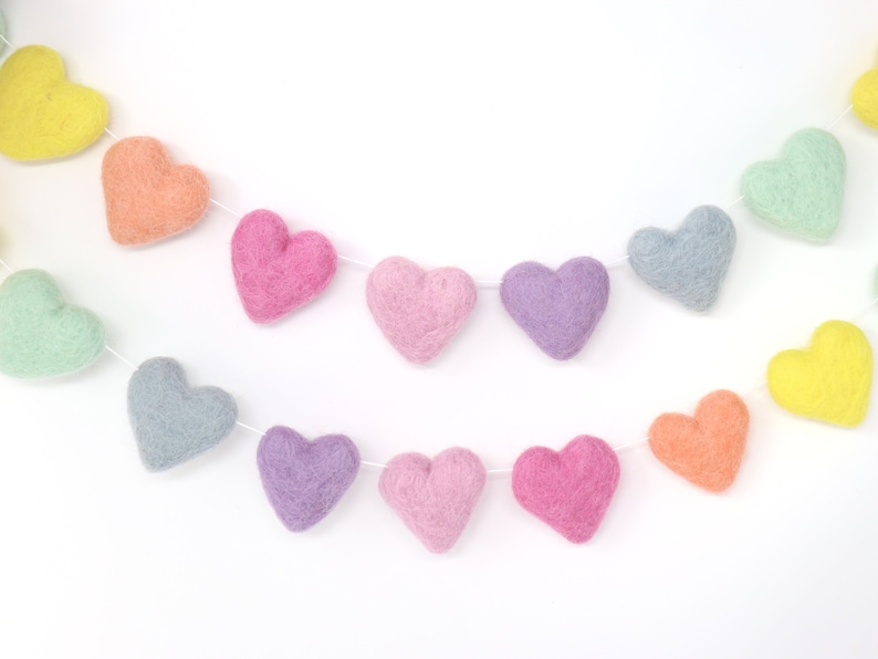May include: A colorful felt heart garland in pastel shades of yellow, orange, pink, purple, and blue. The hearts are strung together on a white string, creating a festive and decorative item.