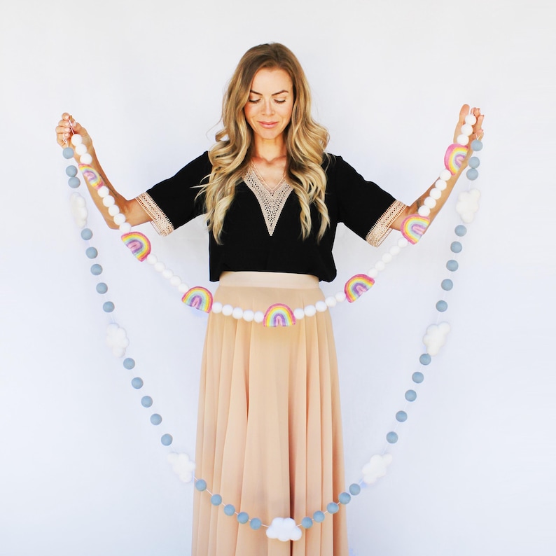 May include: A decorative garland featuring felt rainbows, white clouds, and grey and white felt balls. The rainbow accents have pastel colours. The garland is held up against a white background.