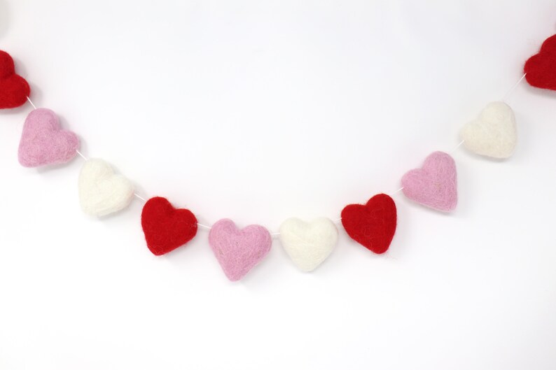 May include: A garland of felt hearts in red, pink, and white. The hearts are strung together with white thread and are arranged in a repeating pattern.