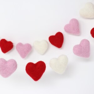 May include: A garland of felt hearts in pink, red, and white. The hearts are strung together with white thread and are hanging against a white background.